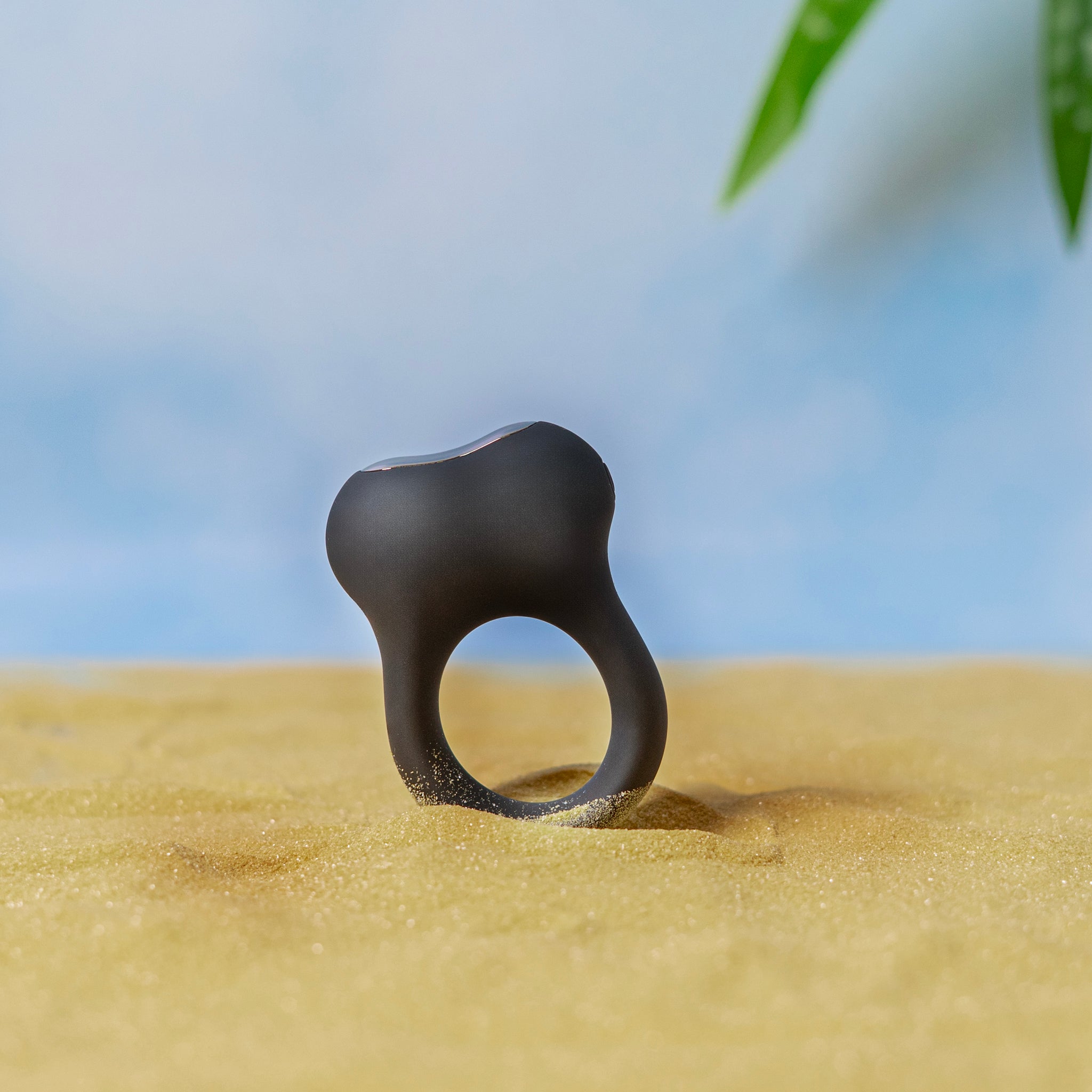 Halo X vibrating cock ring standing upright in sand for a lifestyle-style product photo with a soft blue background.