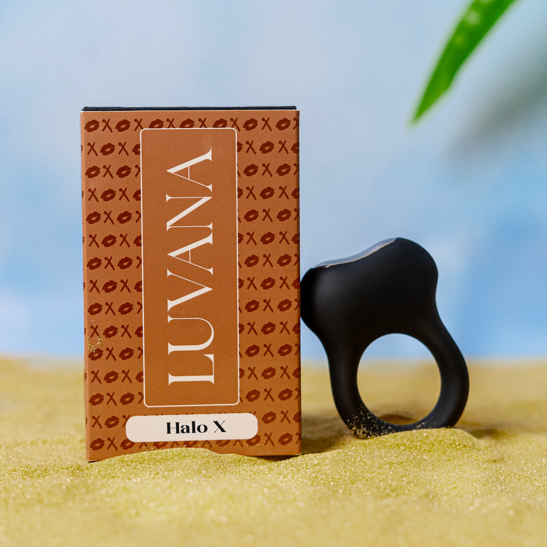 Halo X vibrating cock ring standing upright on soft sand with a blurred blue background for a clean lifestyle product photo.