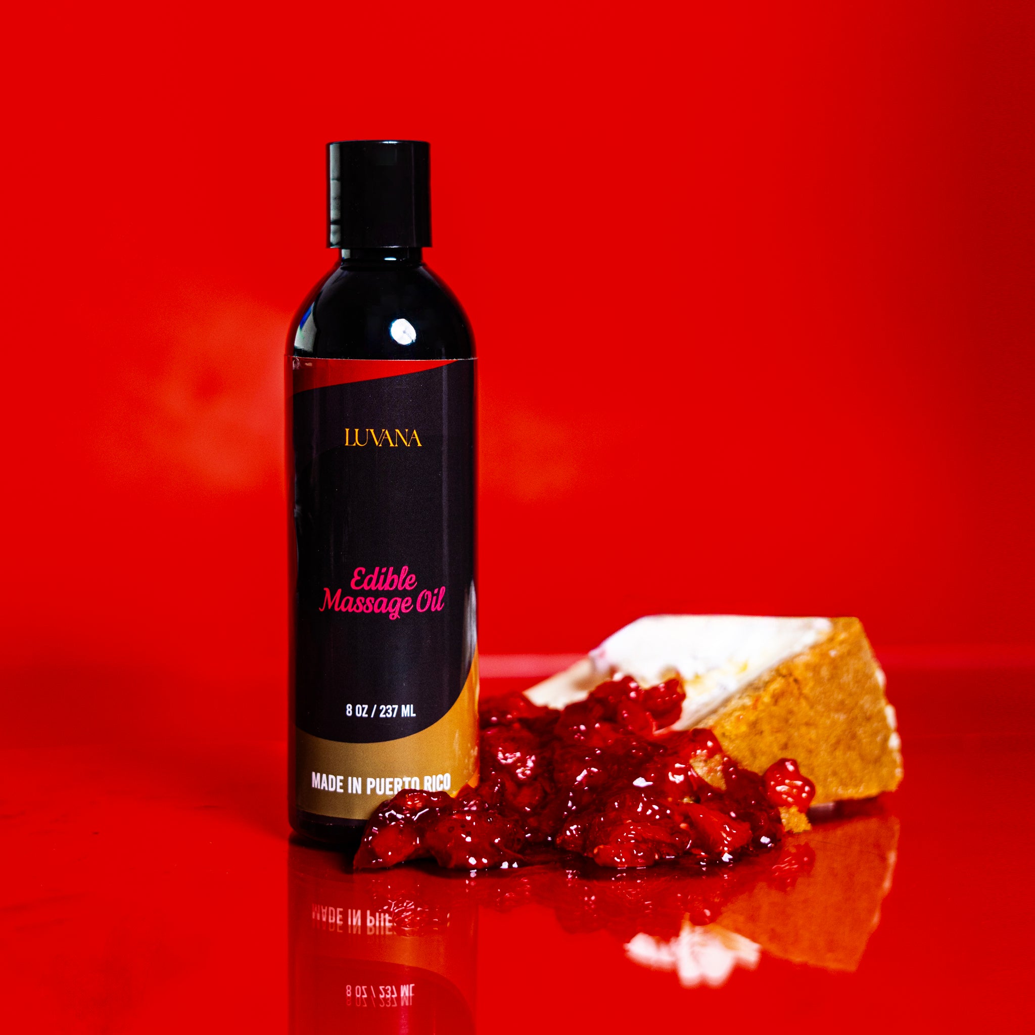 Luvana Edible Massage Oil in Strawberry Cheesecake flavor on a red background next to a slice of cheesecake with berry topping.